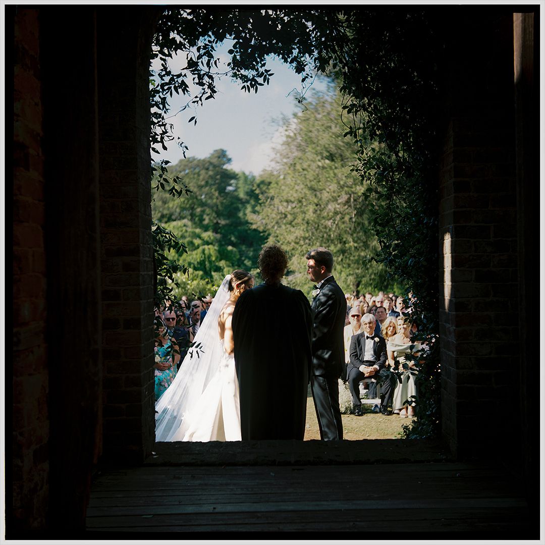Wedding ceremony taking place under an archway with guests seated outdoors in a garden setting.