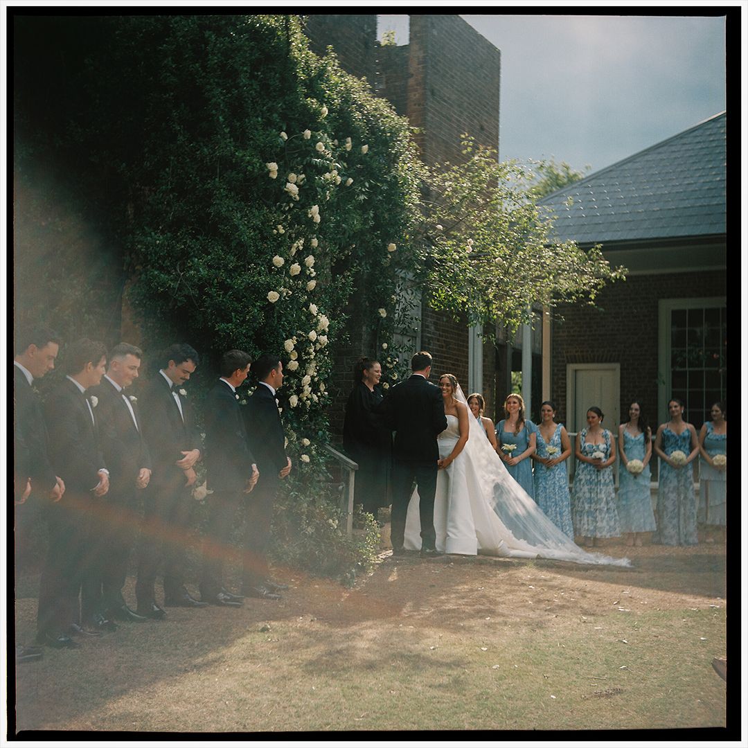 Wedding party stands in line outdoors for a panoramic group photo on a sunny day with trees in the background.