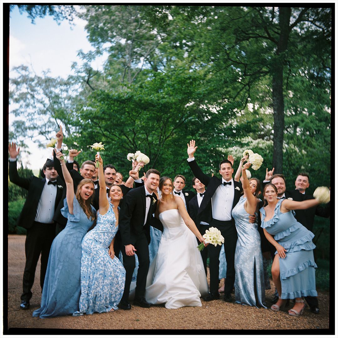Wedding party in blue and floral dresses pose together with newlyweds outdoors in a celebratory group photo.