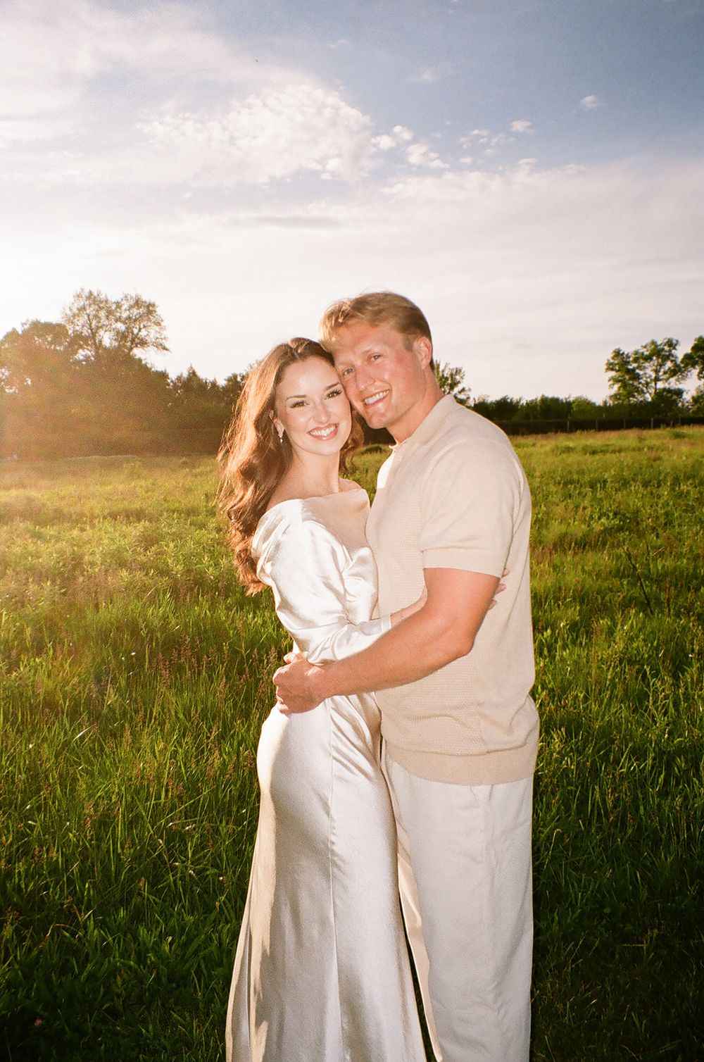 Smiling couple embracing in a sunlit green field during golden hour engagement photo session.