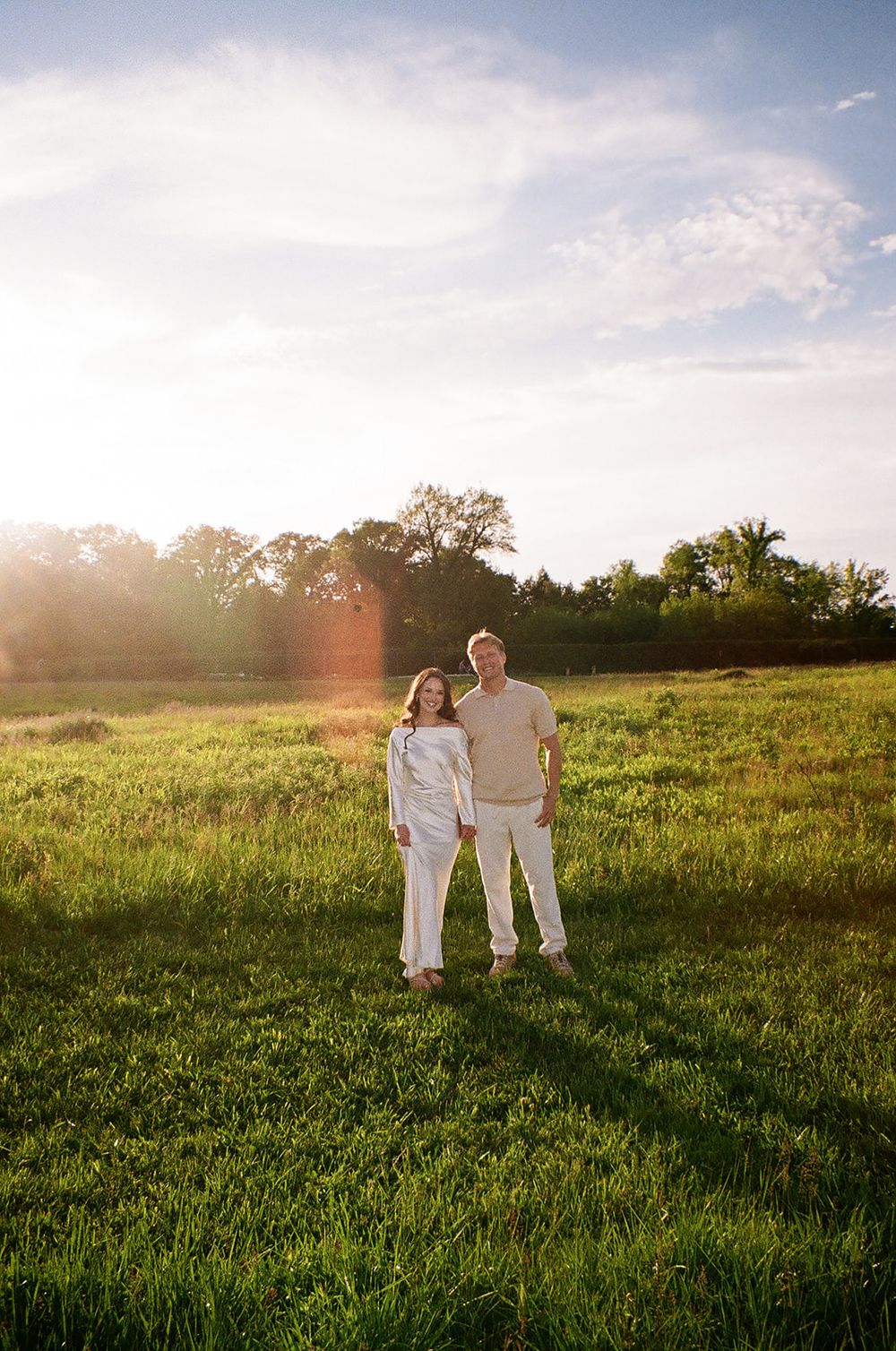 Couple holding hands standing in a vast green field bathed in warm sunset light.