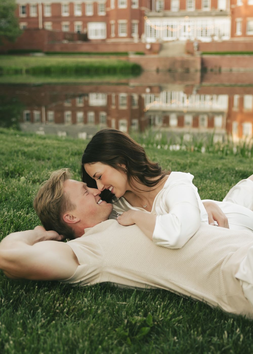 A couple lies on grass near a reflective pond, gazing at each other near a brick building.