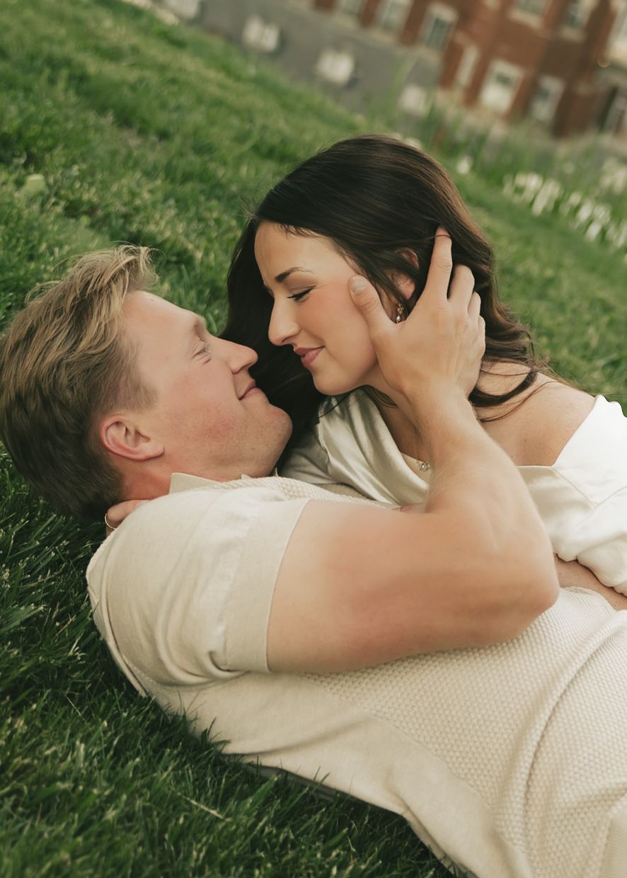 Romantic couple shares intimate moment lying on grass, woman with dark hair cradling mans face tenderly.