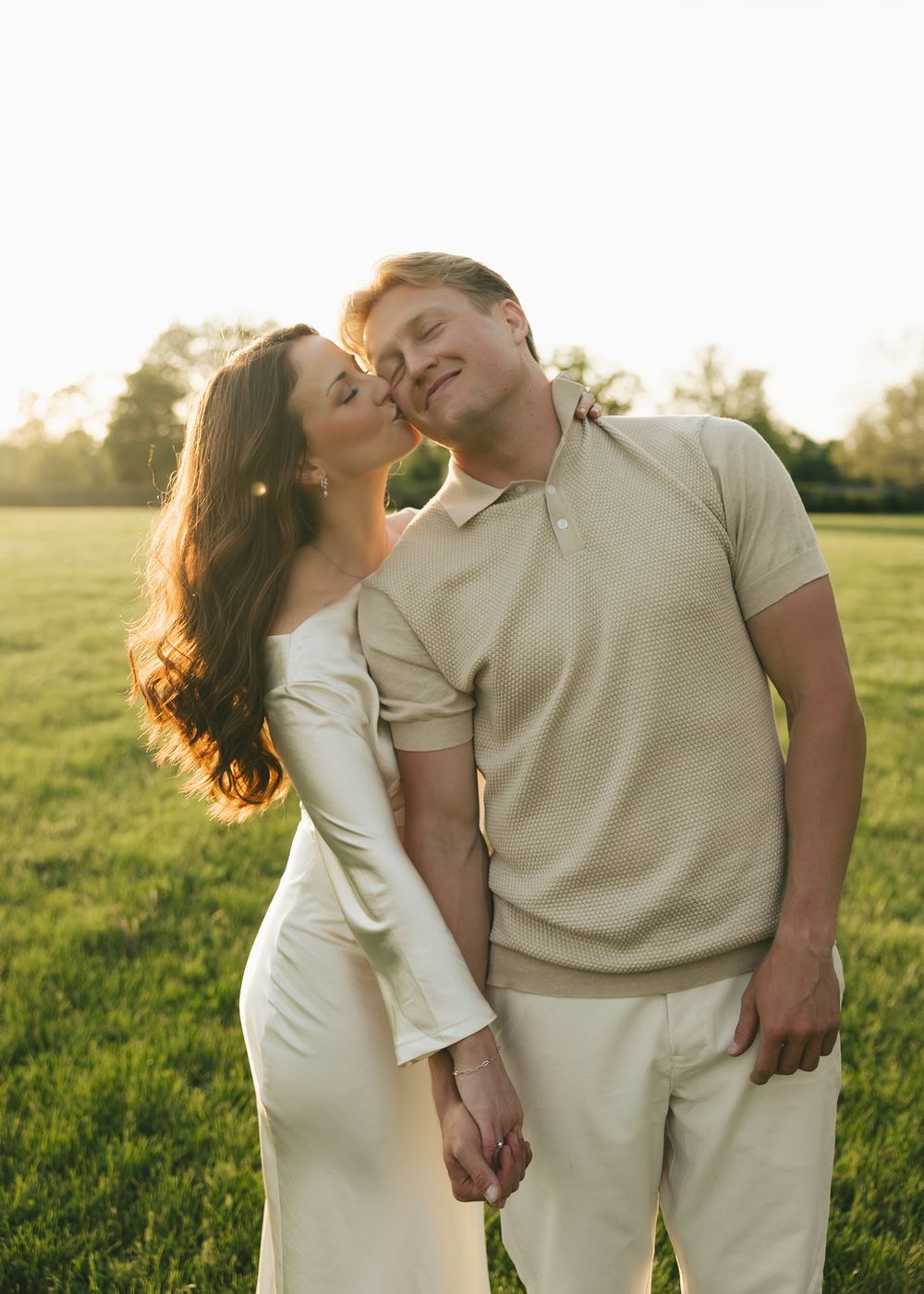 A brunette woman kisses a smiling man on the cheek while holding hands in a green field.