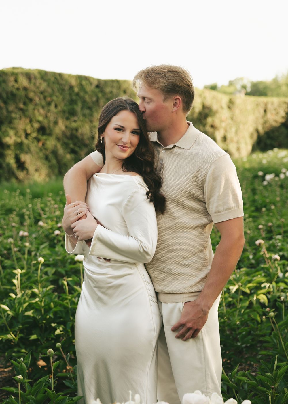 Man kissing woman on the cheek in a garden setting, both wearing coordinating cream and white outfits.