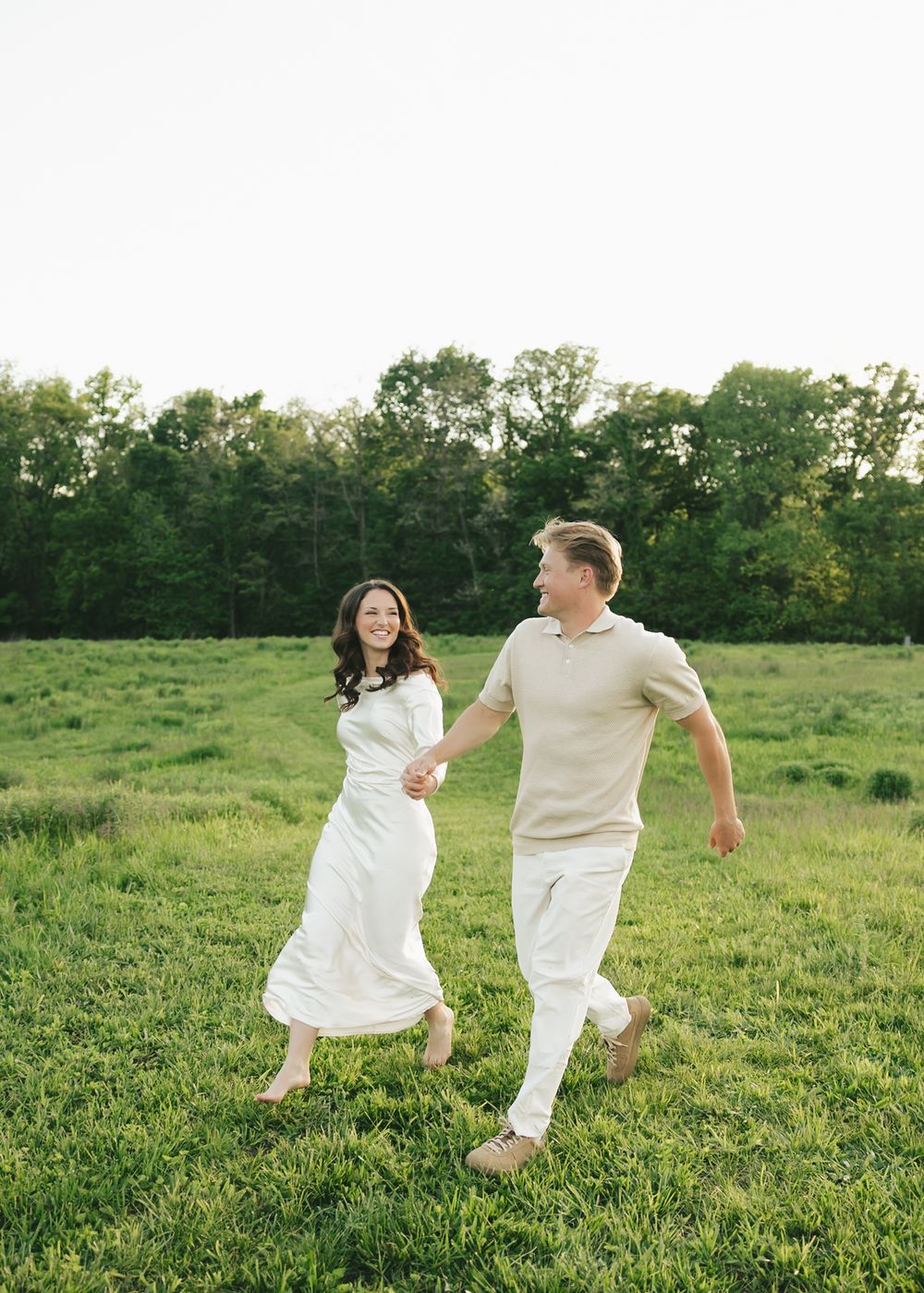 A joyful couple holds hands while walking through a lush green field at golden hour.