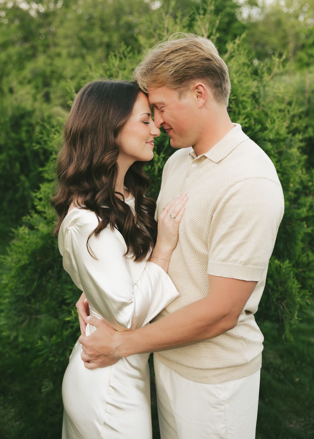 Couple embracing closely in front of lush green hedges during a romantic outdoor engagement photo session.