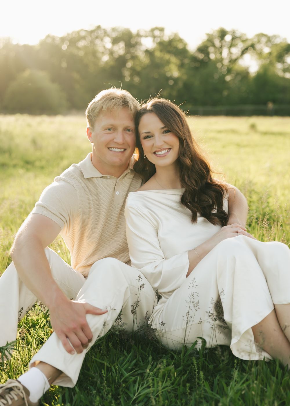 A smiling couple sits together on green grass in a sunlit field, dressed in neutral tones.