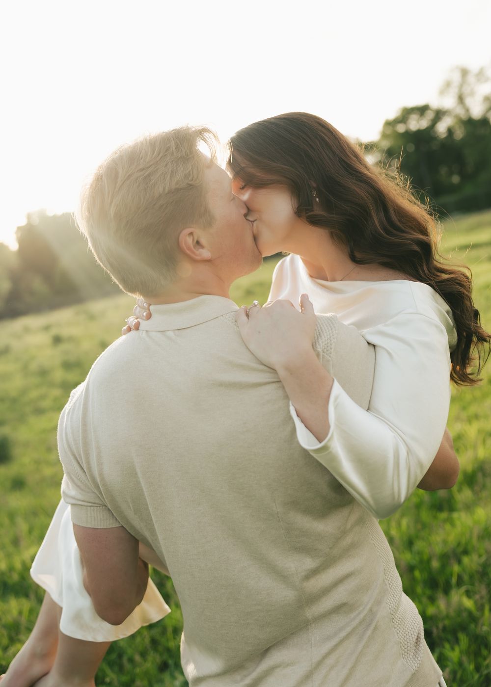 A romantic couple shares a kiss in a sun-drenched green field at golden hour.