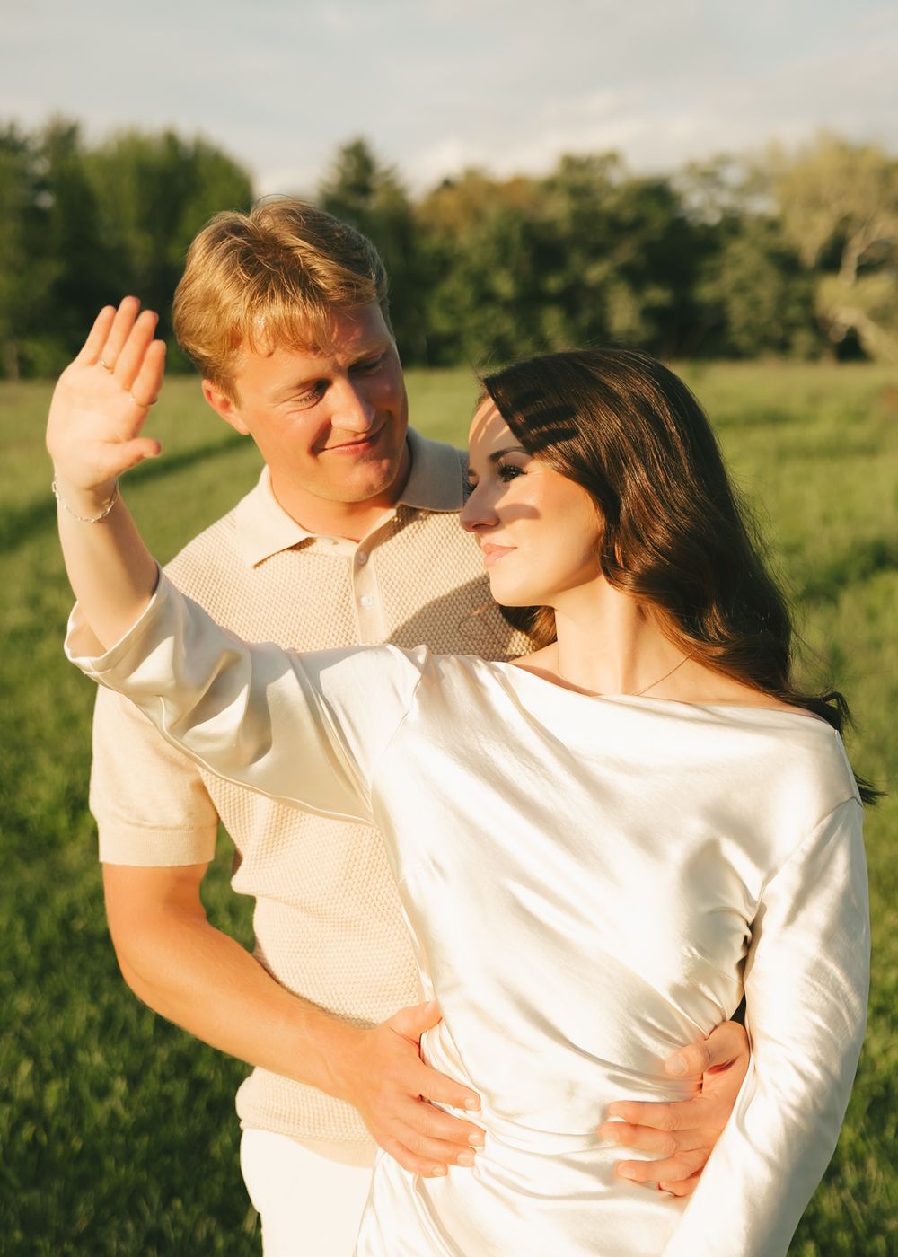 A couple embraces in a glowing sunlit field, the man raising his hand while smiling.