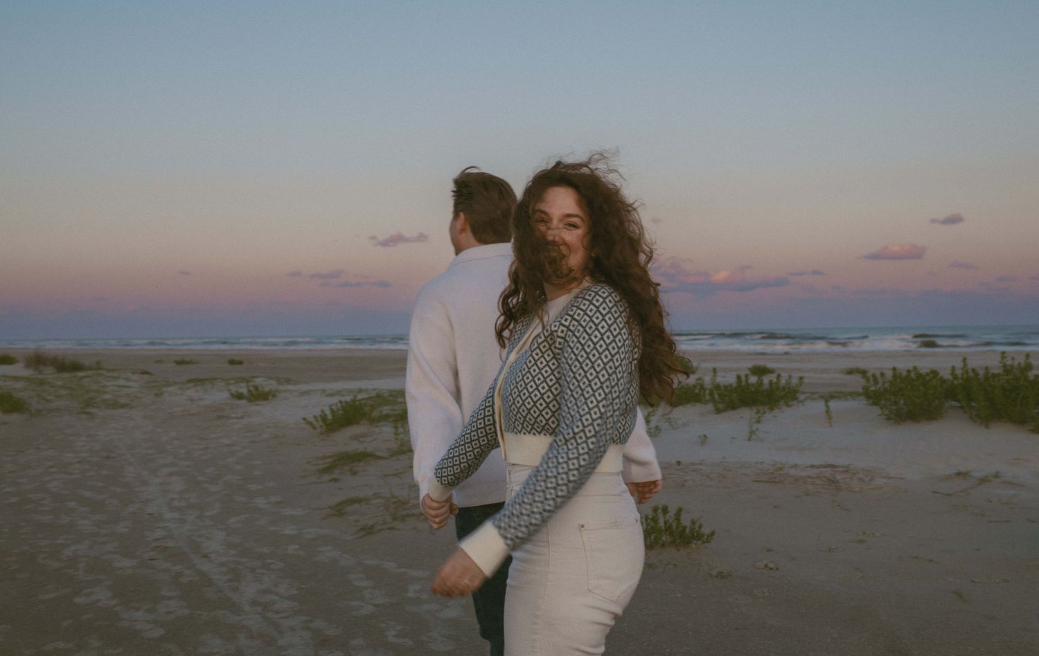 A couple shares a romantic moment for their engagement photos at sunset with soft pink and blue sky in the background.