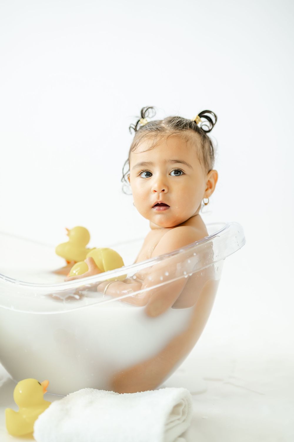Baby girl sitting in a clear bathtub with rubber ducks and bubbles, wearing small earrings with pigtails.