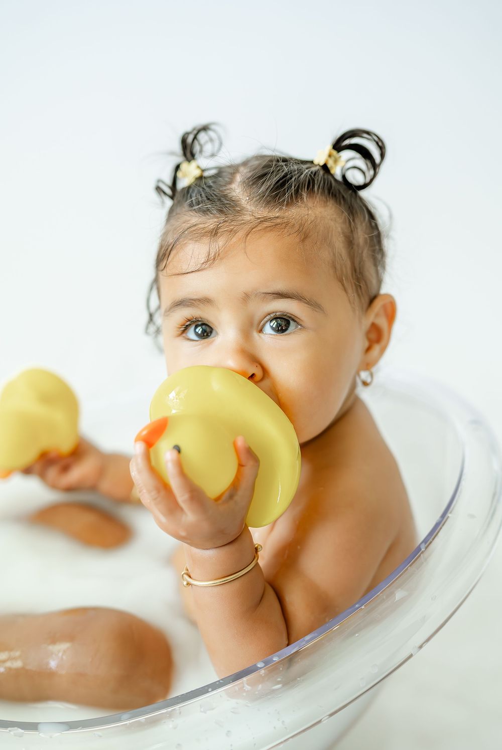 Baby girl in a clear tub chewing on a yellow rubber duck, with pigtails and a gold bracelet.