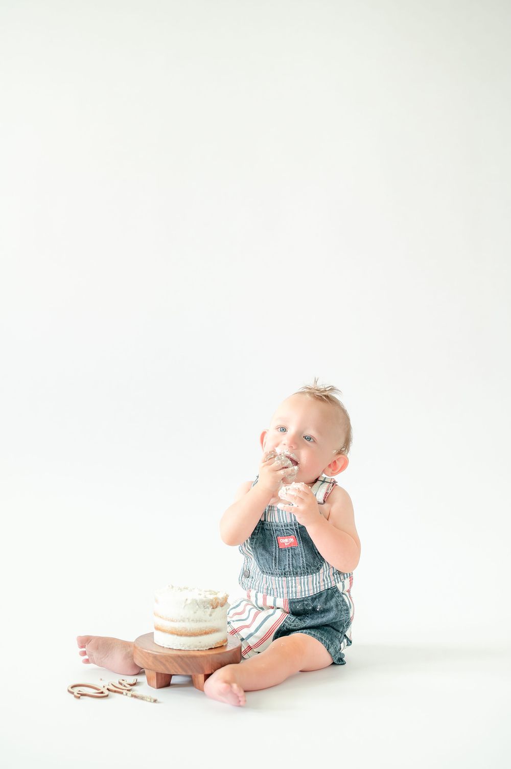 Baby boy in denim overalls eating cake during first birthday cake smash photo session on white background.