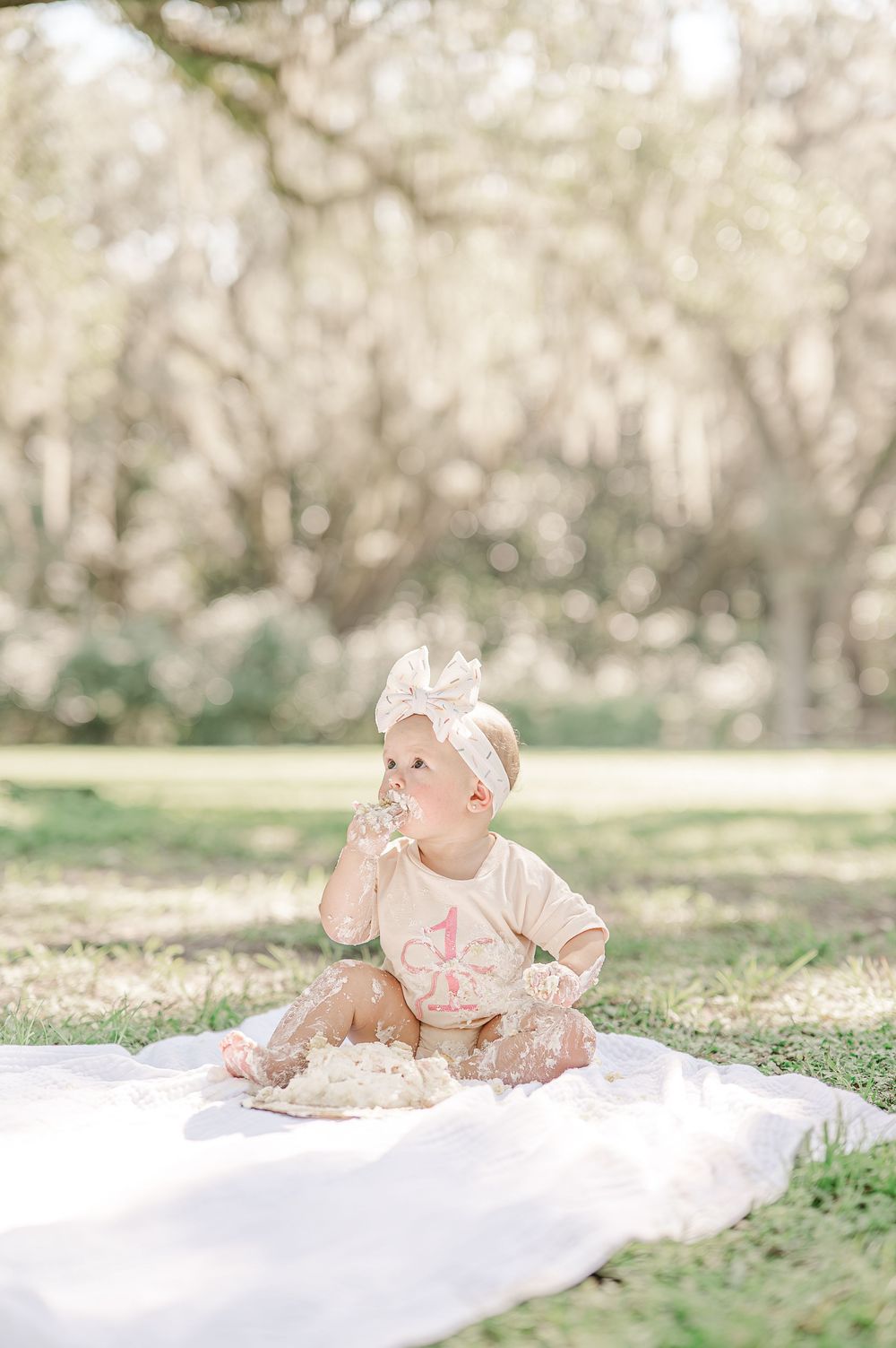 A baby girl in a cream outfit and bow headband sits on a white blanket outdoors, celebrating her first birthday.