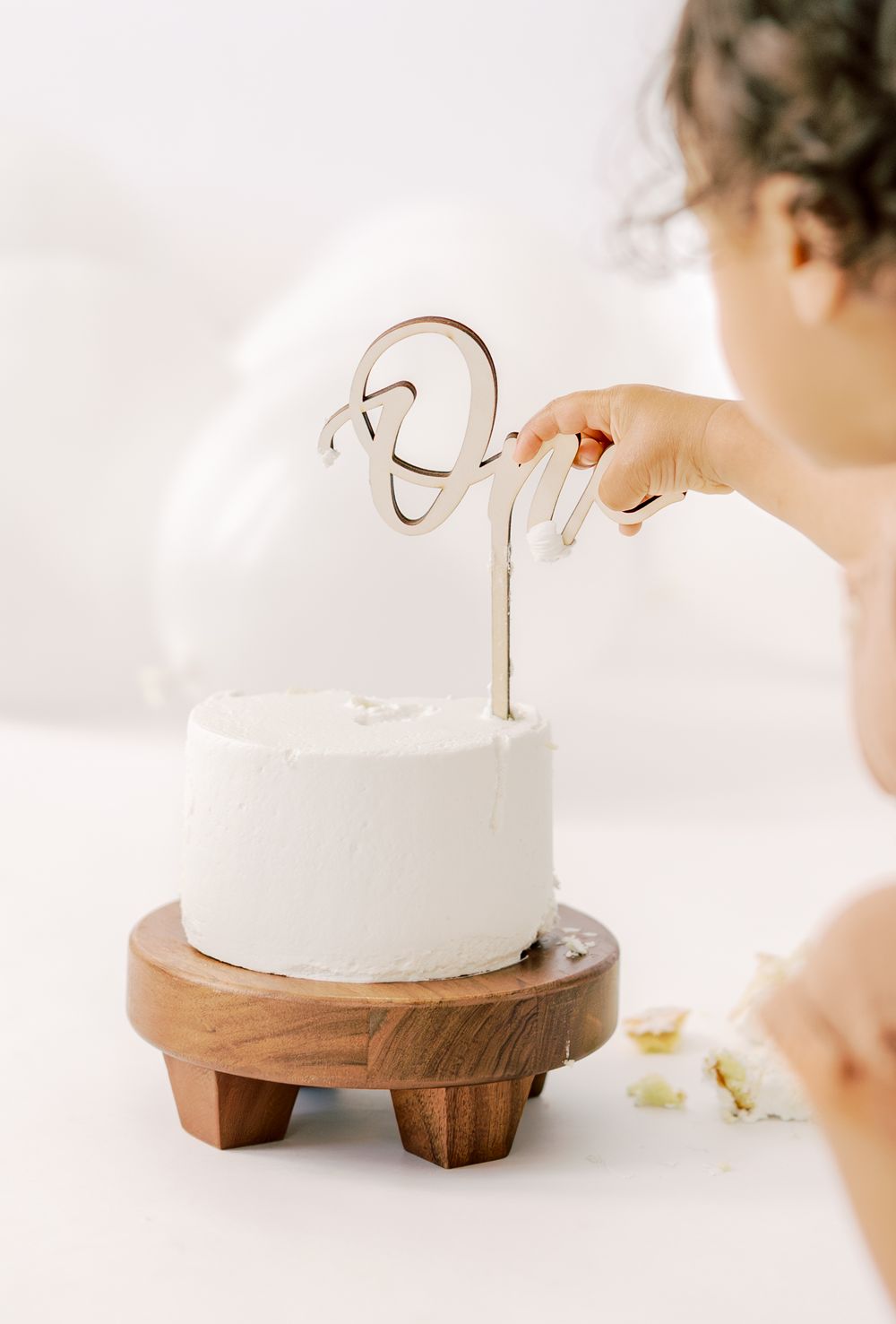 A toddler reaching for a white frosted cake on a wooden stand with a decorative number one cake topper.
