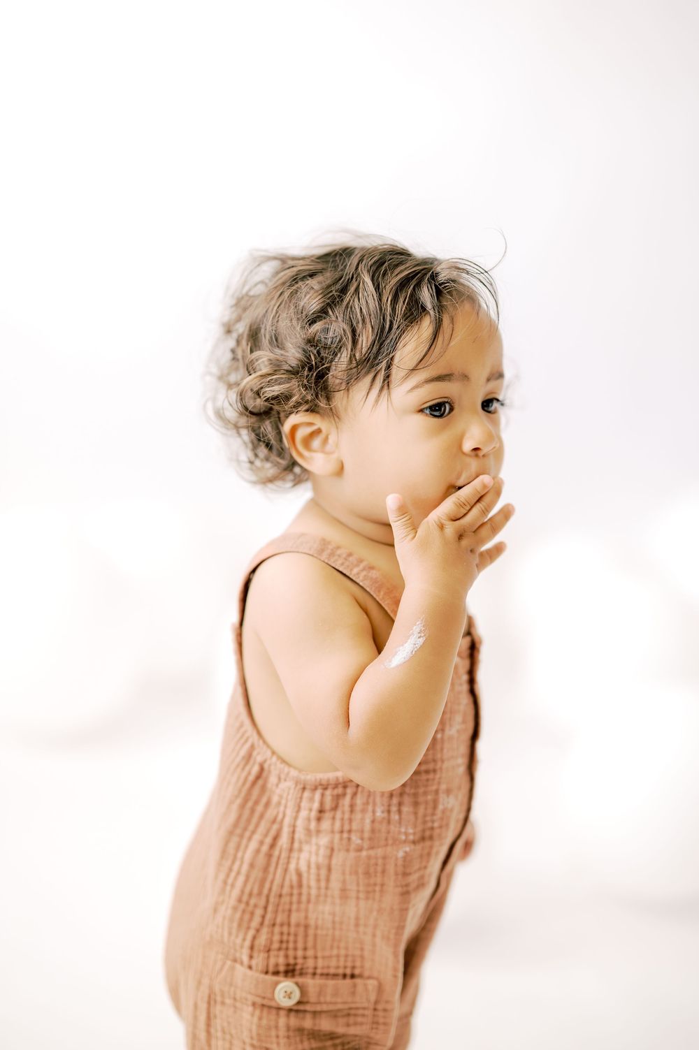Adorable toddler in a brown linen romper with curly hair, hand raised to mouth, against white background.
