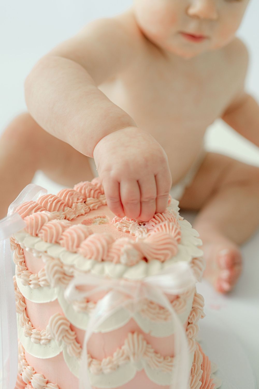Baby reaching into a pink and white frosted smash cake during a first birthday photo session.