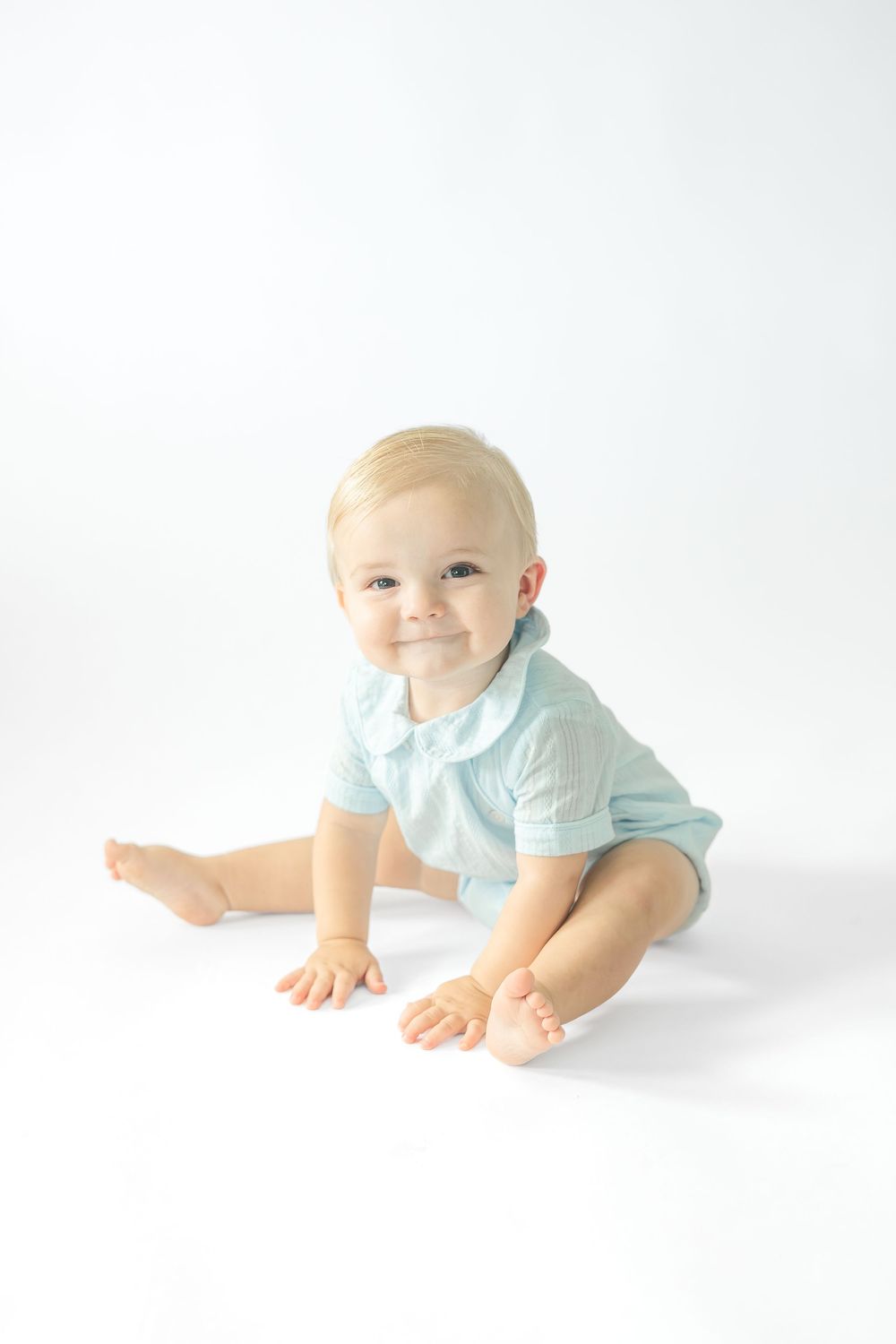 Smiling baby in light blue outfit sitting on white background, looking at camera with a cheerful expression.