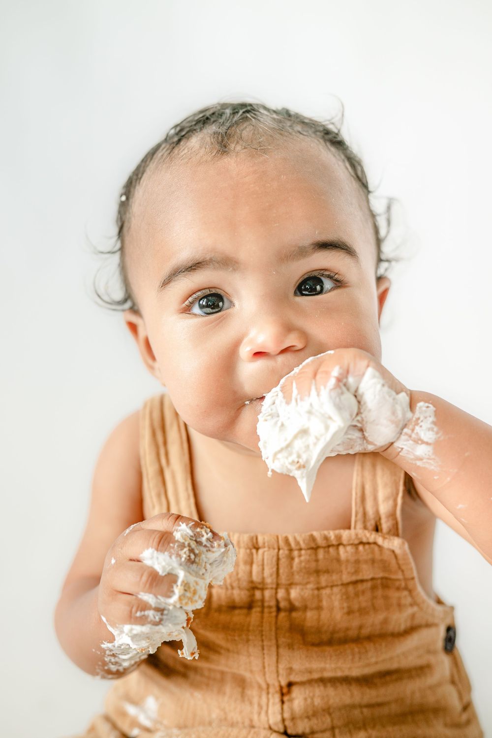 Baby in tan overalls eating white cake frosting with messy hands during a first birthday cake smash photo session.