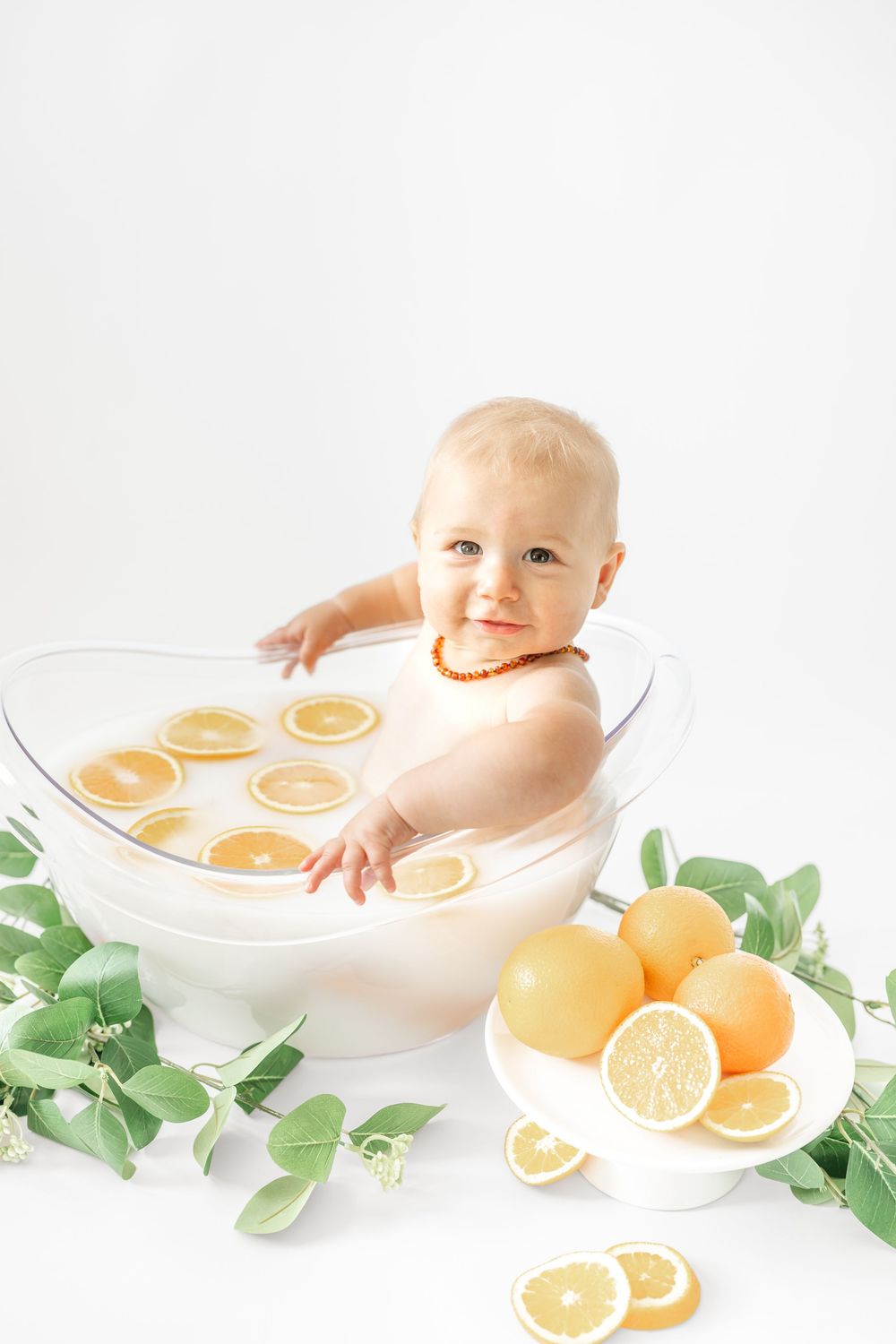 Smiling baby sitting in a clear bowl with lemon slices, surrounded by citrus fruits and green leaves on white background.