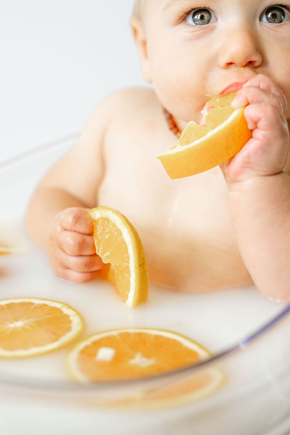 Cute baby with blue eyes tasting a fresh orange slice while sitting in a milk bath with floating orange slices.