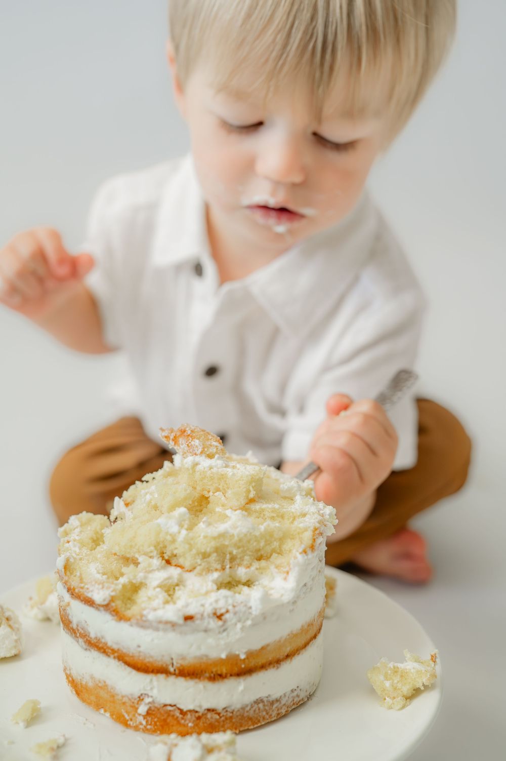 Toddler boy in white shirt smashing a layered white frosted cake during a first birthday cake smash photo session.