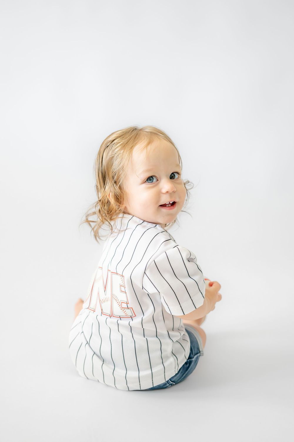 Smiling toddler girl sitting on white background wearing striped baseball jersey and denim shorts, looking over shoulder.