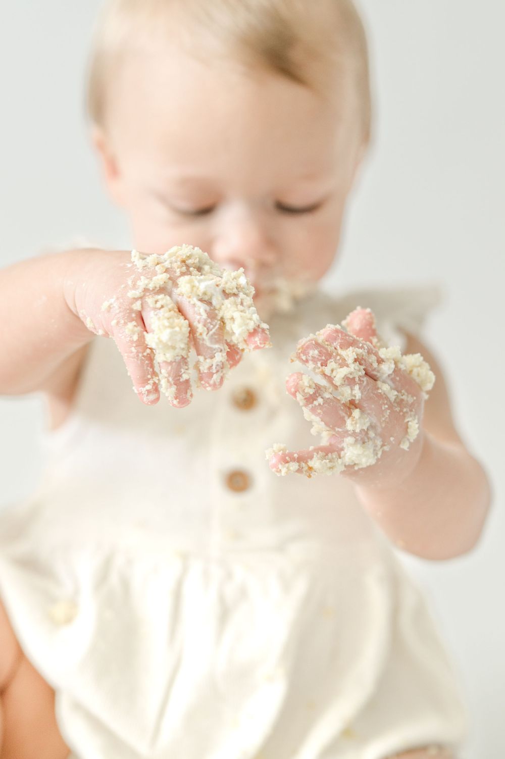 Baby in cream romper holds up cake-smash covered hands during first birthday photo session.