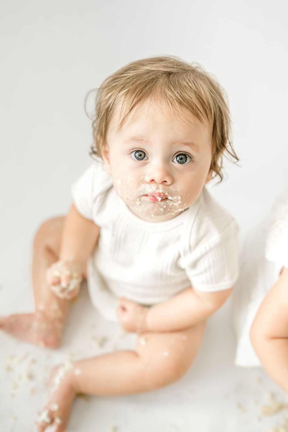 Messy baby with blue eyes in white onesie sits on floor with cake smeared on face during first birthday smash.