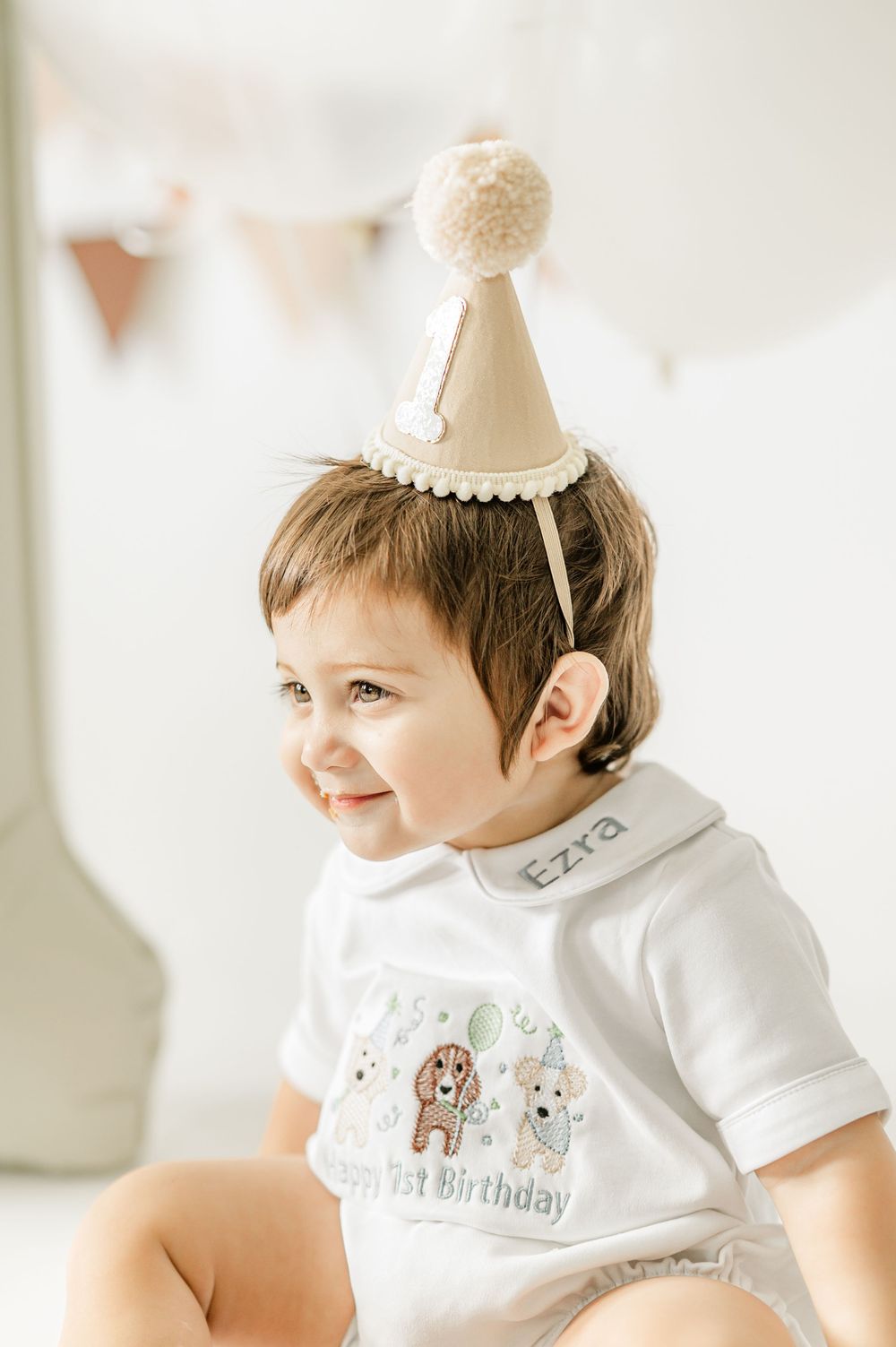 Smiling baby boy wearing a beige party hat and personalized Ezra birthday onesie celebrating his 1st birthday.