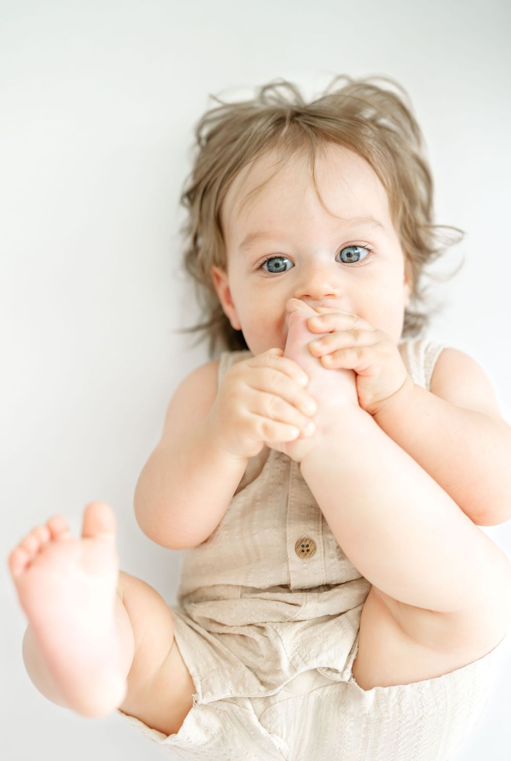 Adorable baby with blue eyes and curly hair sitting up, wearing a linen romper, hands near mouth.