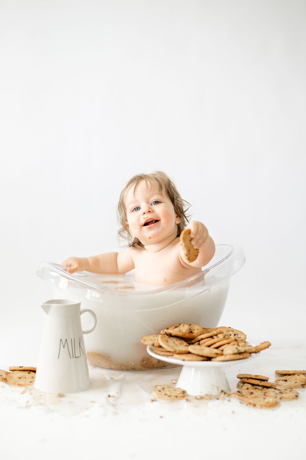 A happy toddler sits in a milk bath holding a cookie, surrounded by crackers and a white milk pitcher.