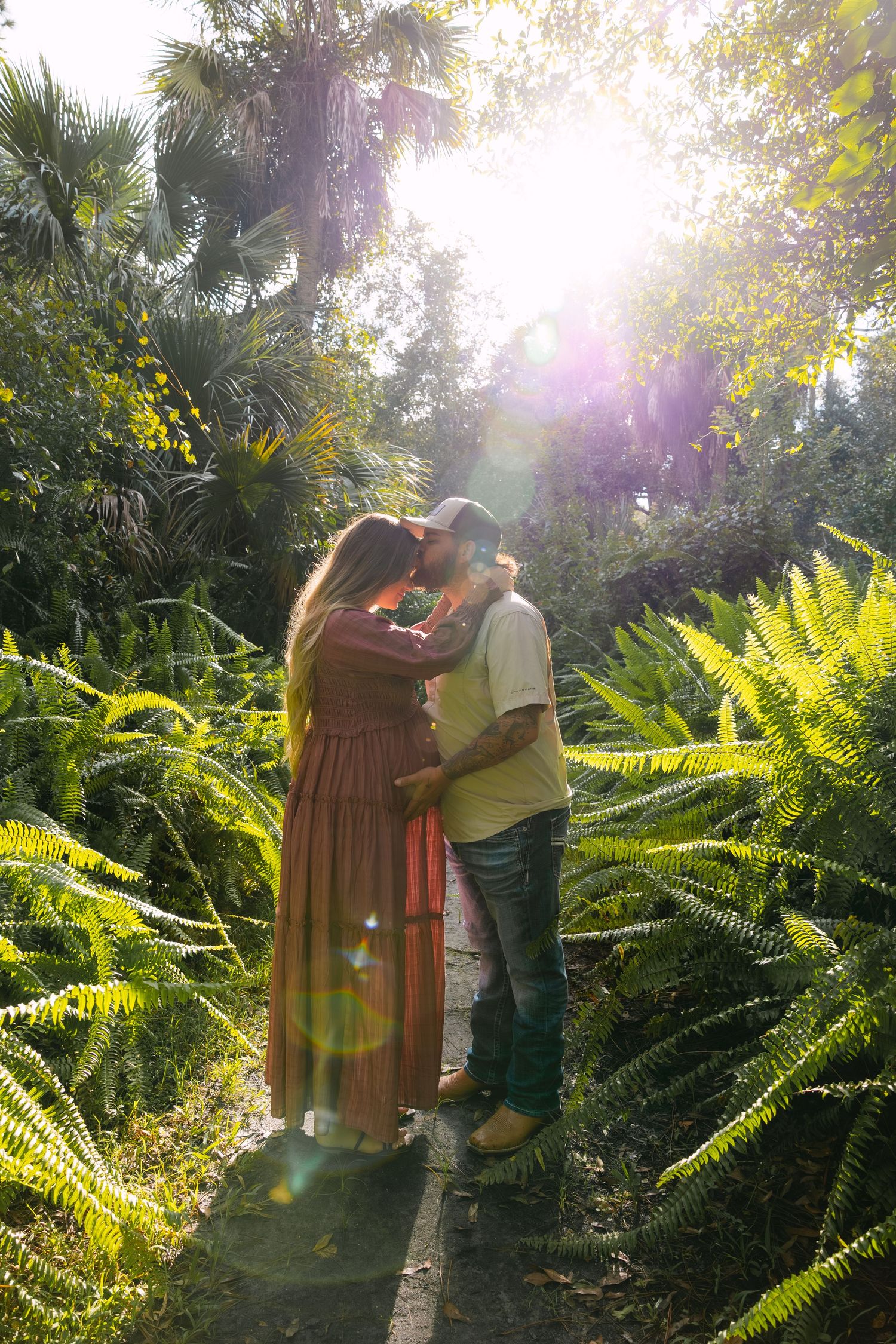 Couple embracing on a sunlit garden path surrounded by lush ferns and palm trees in golden light.