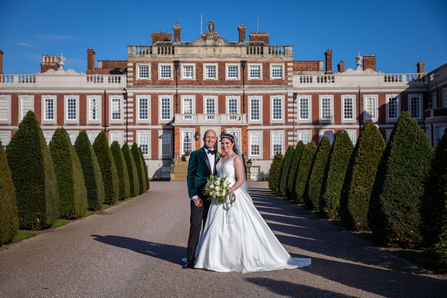 Bride and groom pose together in front of a grand historic manor house surrounded by manicured hedges on a sunny day.
