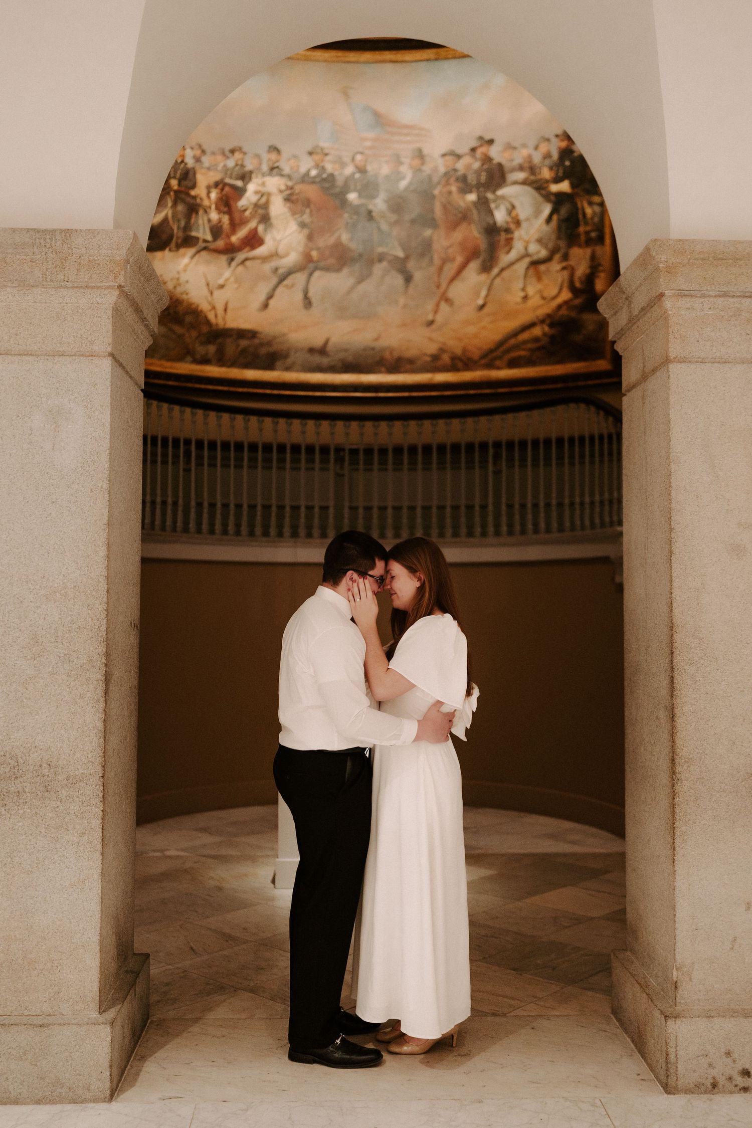 Couple embraces tenderly beneath a domed fresco of a cavalry battle scene in a neoclassical building.