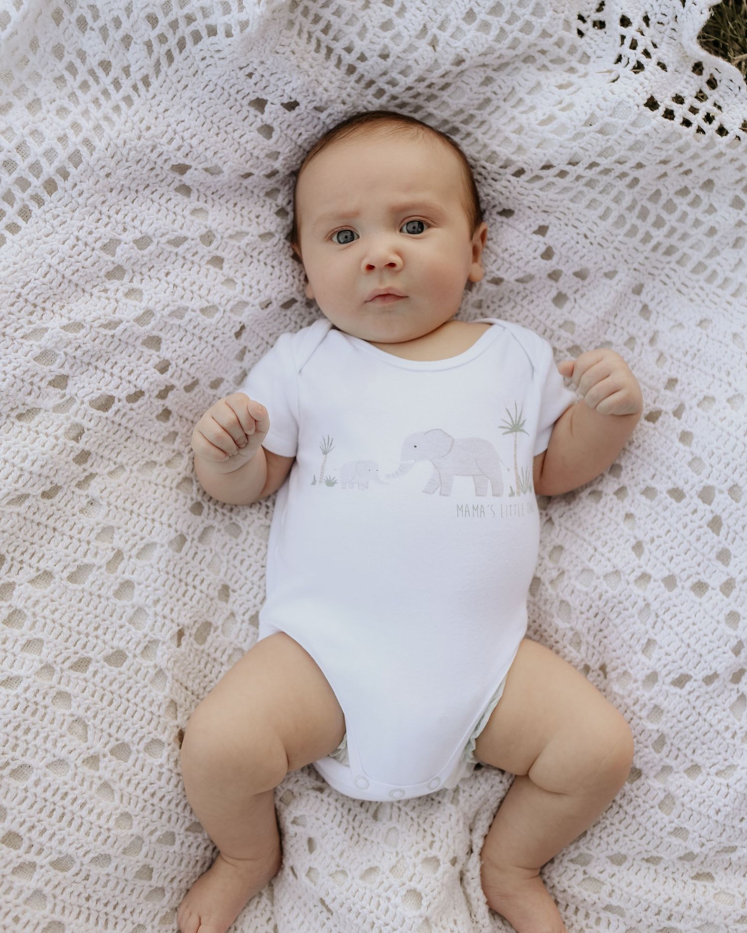 Baby wearing white onesie with elephant design lying on delicate lace blanket.