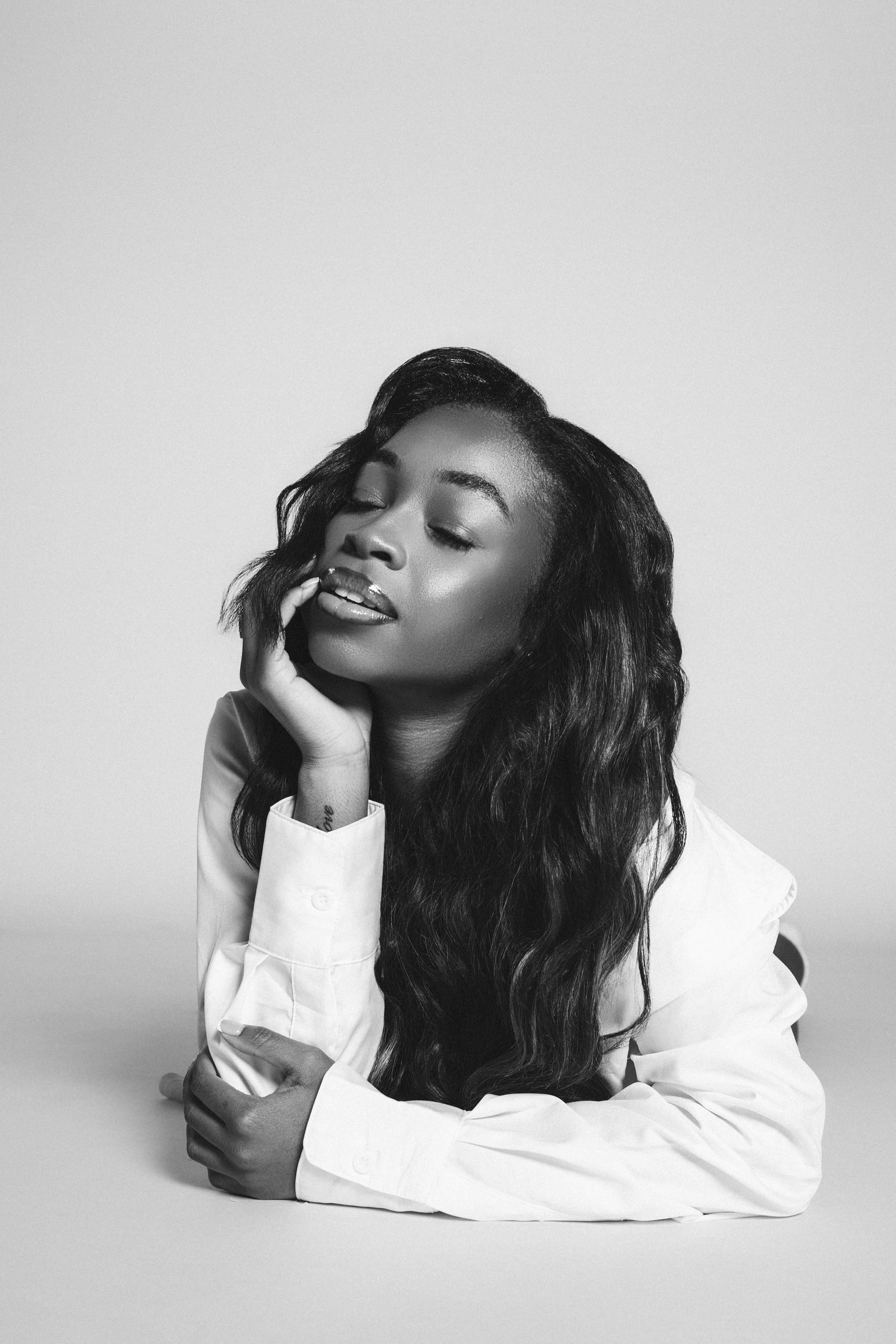 Artistic black and white portrait of a person with long wavy hair in a white shirt posing with eyes closed and hand on cheek.