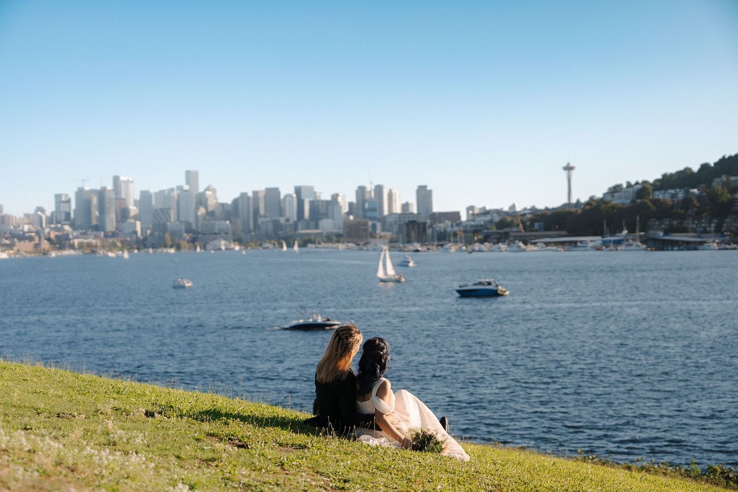 Panoramic view of Seattle skyline and waterfront with boats on Lake Union and people relaxing on grassy hill.