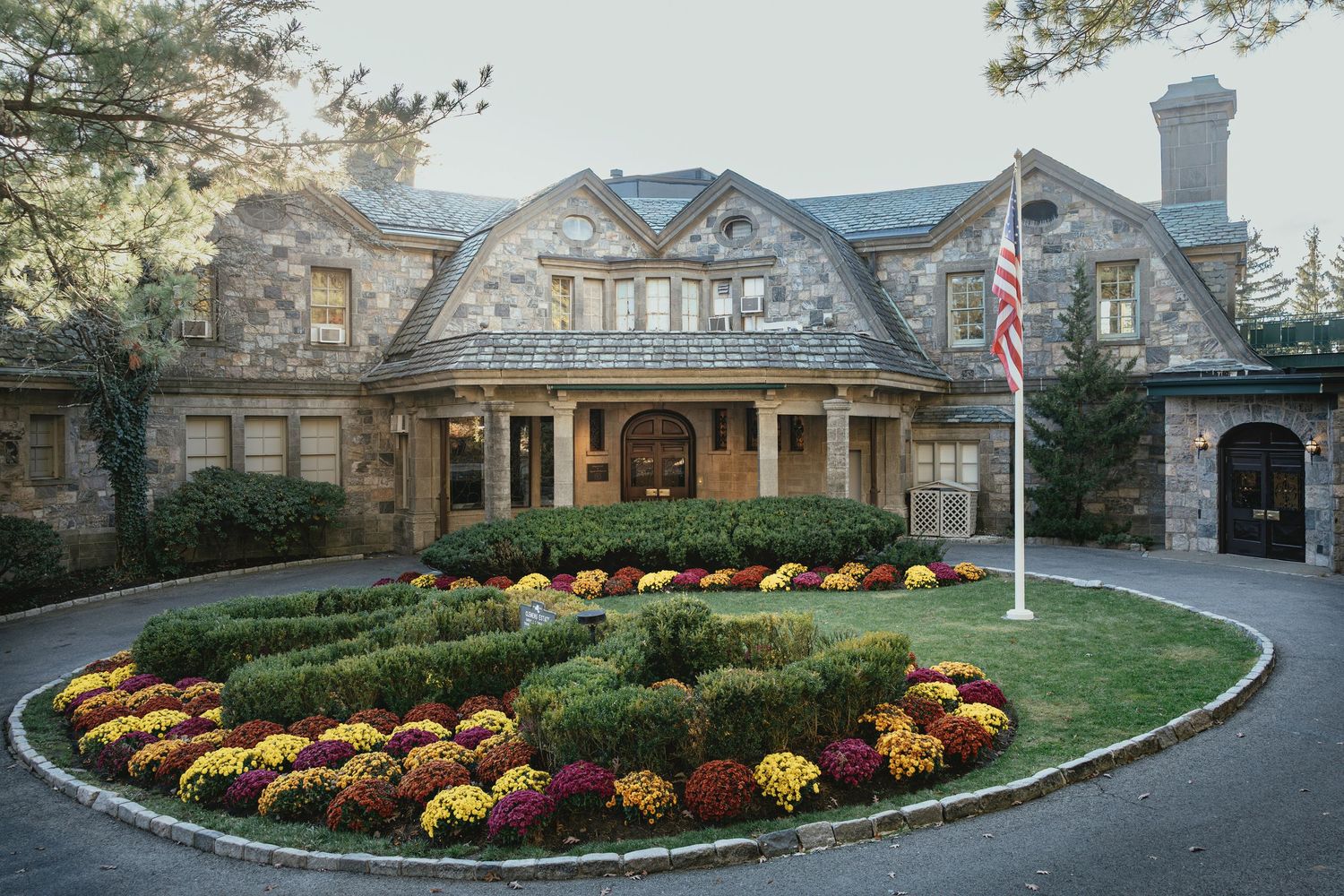 Stone mansion with colorful fall mums planted in curved garden beds along circular driveway entrance.