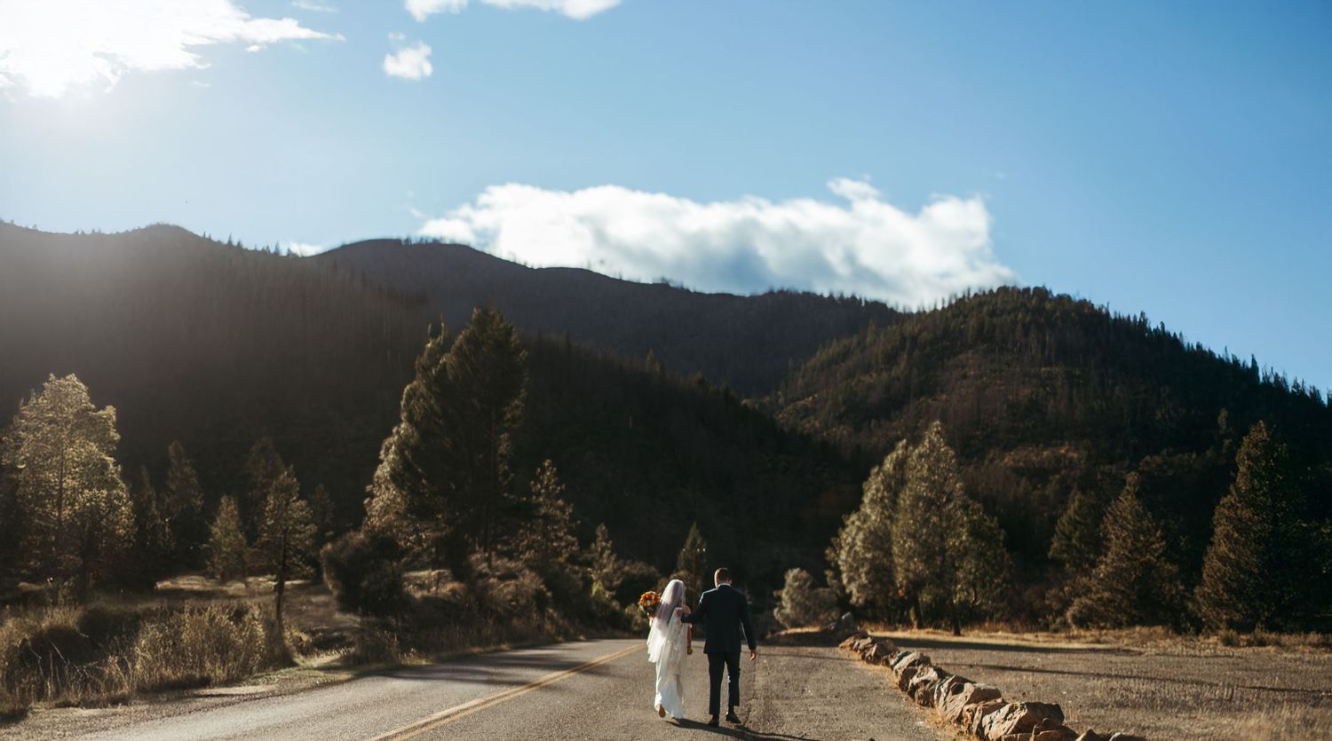 Couple walking hand-in-hand down a mountain road surrounded by pine trees and blue skies in a scenic landscape.