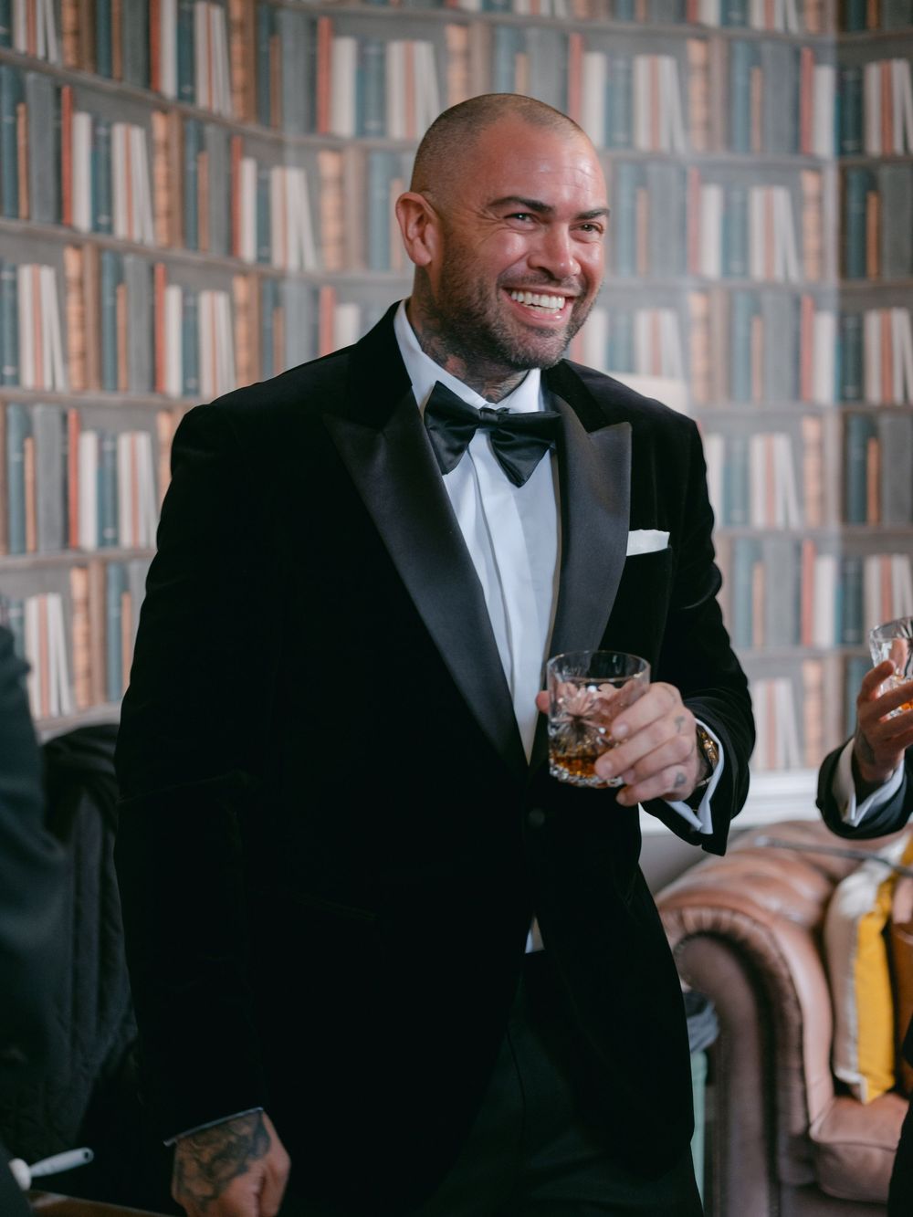 A man in a black tuxedo and bow tie laughs while holding a glass of whiskey at an elegant event.