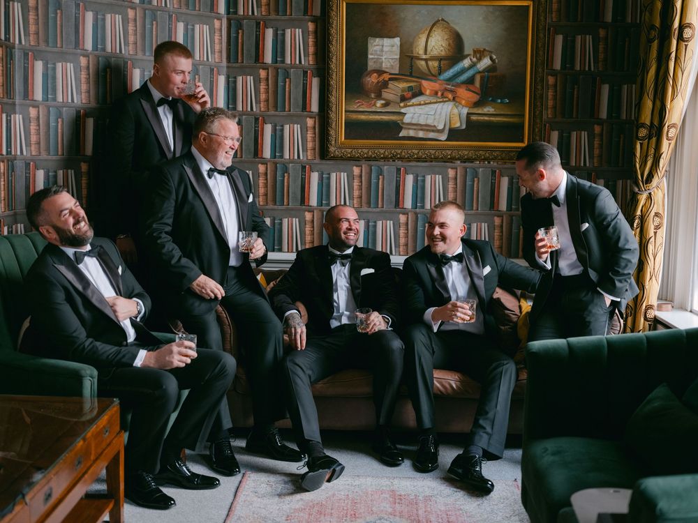 Four people in black tuxedos sit on a leather couch in an elegant library setting.