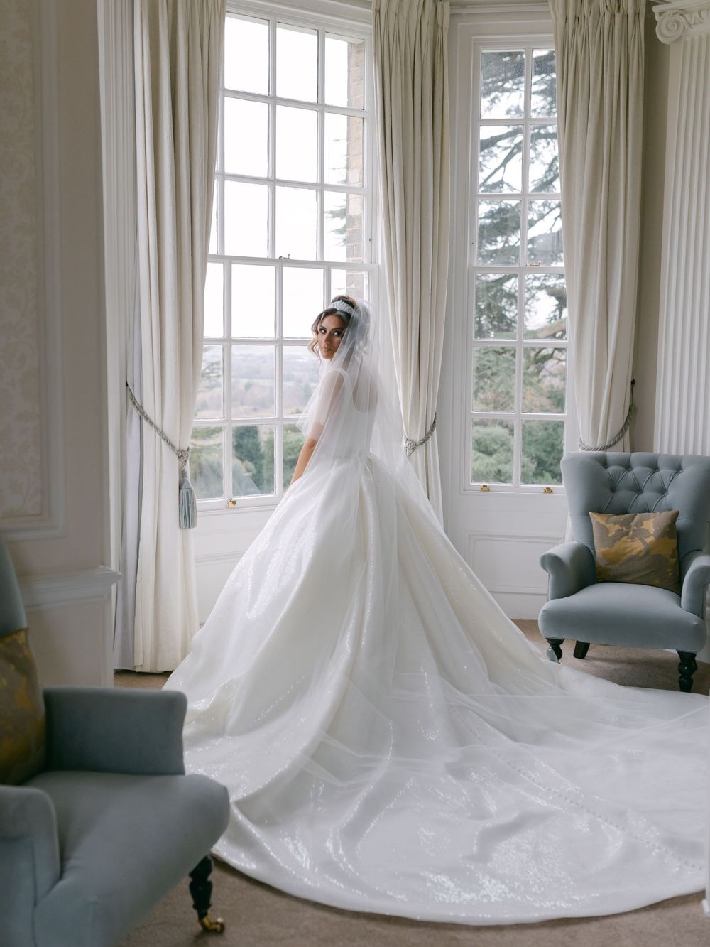Elegant white wedding gown with dramatic cathedral train photographed in a bright sunlit room with blue velvet chairs.
