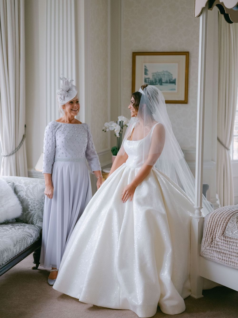 Two people in formal attire share a moment in an elegant room, one wearing a blue-grey dress and one in a white wedding gown.