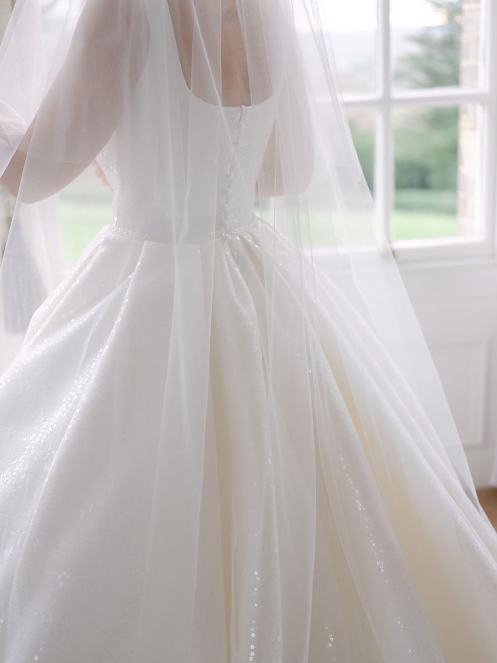 Close-up detail of a white wedding dress bodice with delicate veil and sparkly embellishments near a bright window.