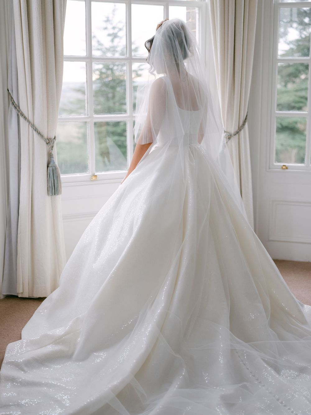 Elegant white ball gown wedding dress with flowing train and veil photographed from behind in a sunlit room.