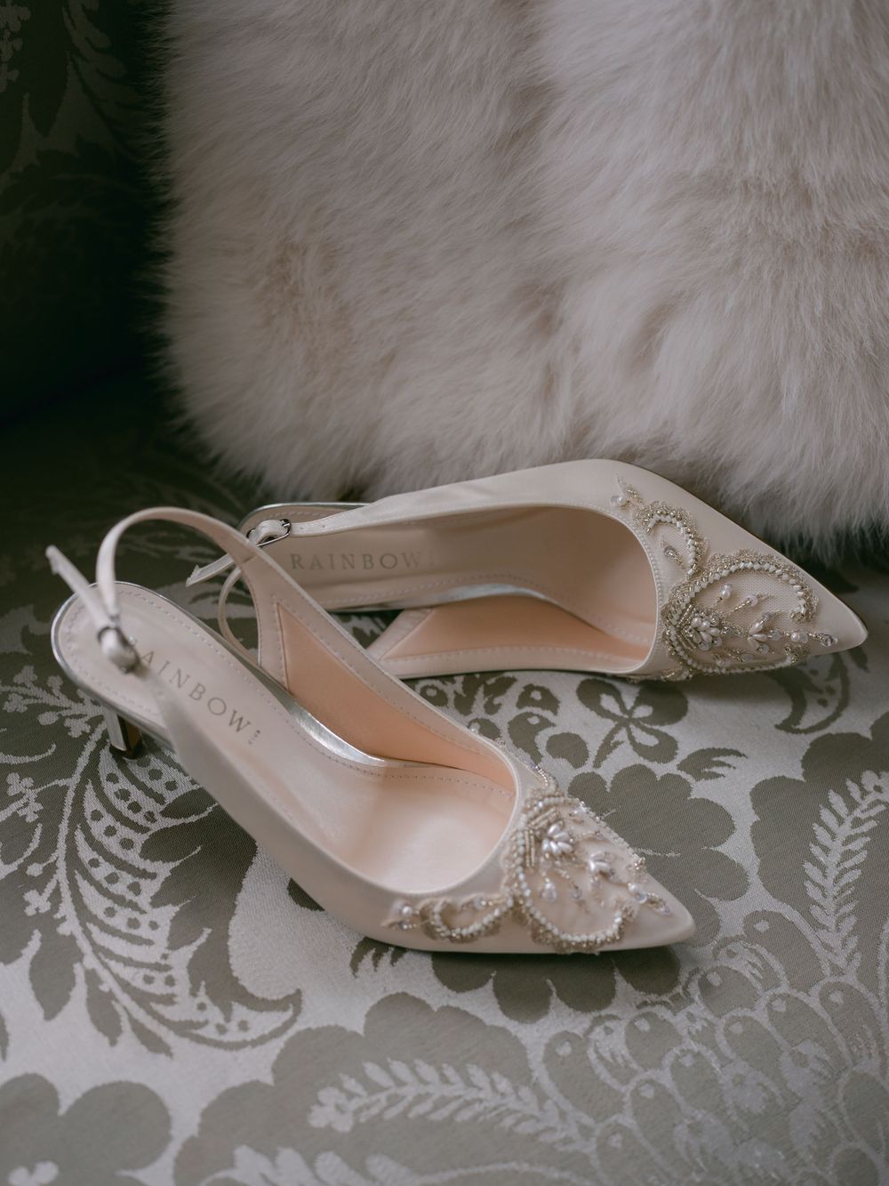 Elegant white lace slingback bridal shoes with pointed toes displayed on a damask patterned fabric.