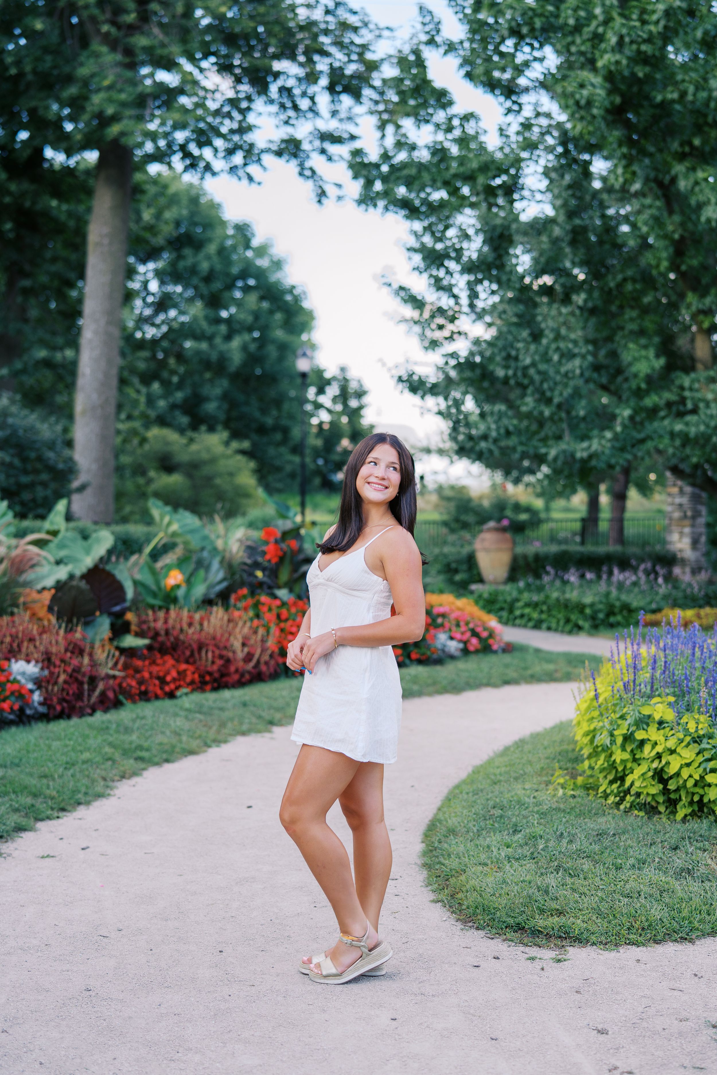 Person in white dress poses on garden path with colorful flower beds and trees.