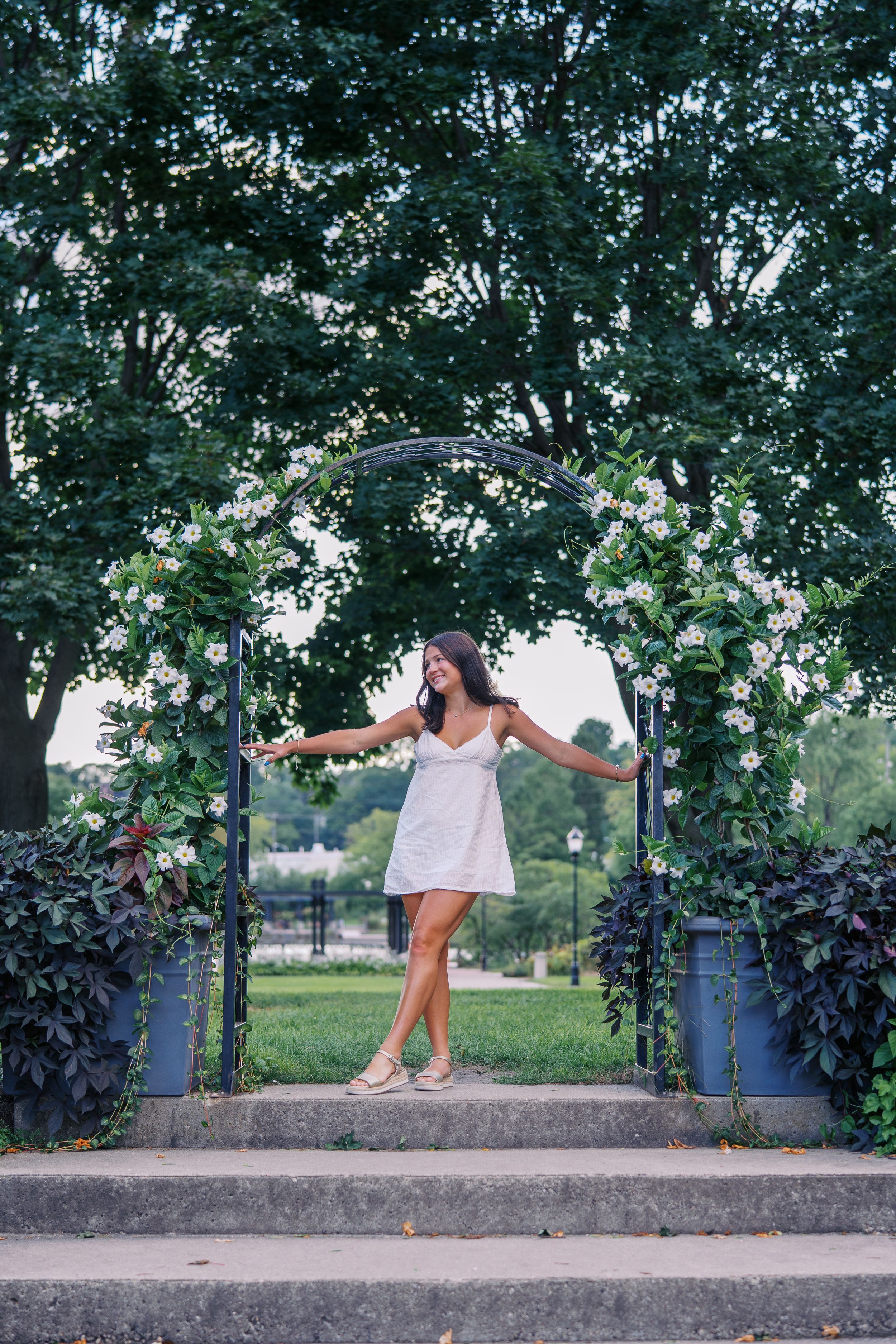 Person stands under floral arch on stone steps with arms outstretched in joyful pose.